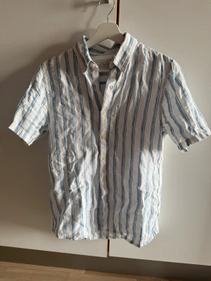 Linneskjorta - Fräsch kortärmad skjorta från Abercrombie & Fitch vitt med blå ränder. Skjortan är tillverkad i 100% linne och har klassisk krage samt knäppning framtill. Perfekt för en avslappnad och stilren look.