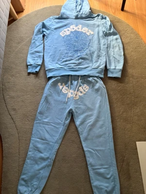 Blått loungewear-set från Sp5der - Säljer ett blått loungewear-set från Sp5der med hoodie och matchande byxor. Hoodien har tryckt spindelnätsmotiv och logga framtill, samt känguruficka och huva. Byxorna har resår i midjan med dragsko och tryckt logga. Perfekt för chill dagar hemma.