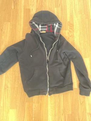 Svart hoodie med rutigt foder - Snygg svart hoodie med dragkedja och huva. Insidan av huvan och öppningen har ett klassiskt rutigt mönster i svart, vitt och rött. Två fickor framtill och justerbara snören i huvan. Perfekt för en chill och avslappnad stil.
