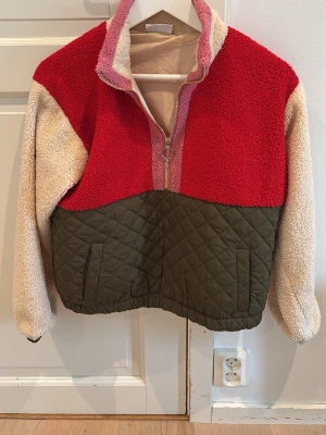 Färgglad teddyjacka med quiltad nederdel - Teddyjacka i röd, beige och rosa med quiltad olivgrön nederdel. Jackan har halv dragkedja med ringdetalj, hög krage och två fickor framtill. Materialet är mjuk teddyfleece upptill och quiltad polyester nedtill. Perfekt för dig som gillar statement-plagg.