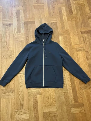Grå hoodie från BOSS - Snygg grå hoodie från BOSS med dragkedja framtill och justerbar huva med snören. Diskret BOSS-logga på bröstet och två fickor framtill. Tillverkad i ett mjukt och bekvämt material som passar perfekt till en avslappnad stil.