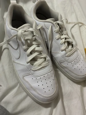 Vita Nike sneakers i läder - Fräscha vita sneakers från Nike med klassisk låg siluett. Skorna är tillverkade i slätt läder med perforerade detaljer på tån och sidorna för extra ventilation. Tjock vit sula och matchande vita skosnören ger en clean look som passar till det mesta.