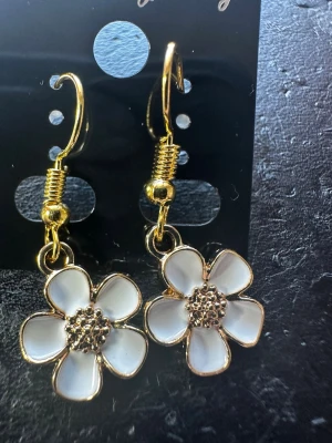 Blommiga örhängen i guld och vitt - Supersöta örhängen med hängande blommor i vit emalj och guldfärgade detaljer. Blommorna har fem kronblad och en glittrig mitt, vilket ger en fräsch och lekfull look. Perfekta för att addera lite extra stil till din outfit.