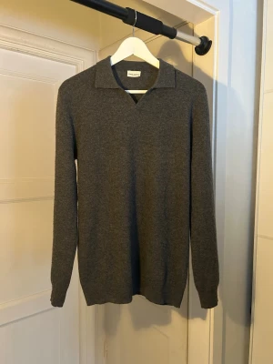 Mörkgrå stickad tröja med krage - Säljer en stilren mörkgrå stickad tröja från Arket. Tröjan har en klassisk krage med v-ringning och långa ärmar. Materialet ser ut att vara mjukt och av hög kvalitet, perfekt för lager på lager under kyliga dagar.