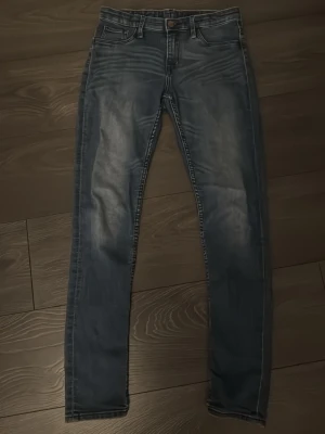 Blå skinny fit jeans - Säljer ett par blå slim fit jeans med klassisk femficksdesign. Jeansen har en knappstängning och dragkedja framtill samt ljusa detaljer på låren och bakfickorna. Perfekt för dig som gillar en modern och avslappnad look. Passformen är som slim fit men heter skinny fit. Finns inga fickor heller.