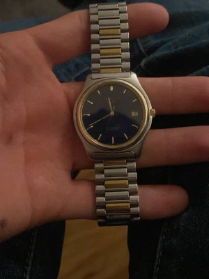 Klassisk Seiko/certina  herrklocka i silver och guld - Stilren Seiko herrklocka med rund urtavla i mörkblått, silverfärgat armband med gulddetaljer och datumvisning. Klockan har en klassisk design med metallarmband och guldfärgad krona. Perfekt accessoar för dig som gillar tidlös stil.    Dena batteri är slut så behövs bytas. Vet bara vad den är värd på ett ungefär.