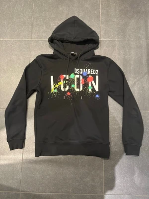 Svart Dsquared2 ICON hoodie - Tjena säljer en Svart hoodie från Dsquared2 med huva och dragsnören. Framsidan har ett färgglatt ICON-tryck med stänk i rött, grönt, blått och gult.
