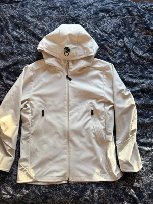 Sail Racing Jacka - Snygg vit vindjacka från sail racing med hel dragkedja, två dragkedjeförsedda fickor och stor huva. Jackan har minimalistisk design och logga på huvan. Perfekt för blåsiga dagar och enkel att matcha med olika outfits. Köptes i november!