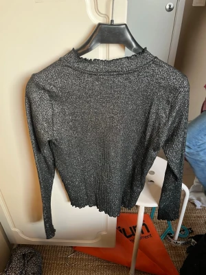 Glittrig svart tröja - Säljer en svart ströja med glittereffekt och lång ärm. Tröjan har ribbad struktur, vågig kant vid hals, ärmslut och nederkant. Perfekt för dig som vill ha något extra till din outfit.