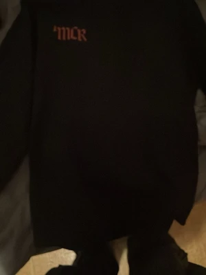 Svart MCR hoodie med tryck - Säljer en svart hoodie med huva från My Chemical Romance. Framsidan har ett stort tryck med bandets namn och en konstnärlig bild i rött och vitt. På bröstet står det 'MCR' i orange text. Perfekt för dig som gillar alternativ stil och bandmerch.