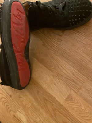 Svarta sneakers med nitar från Louboutin - Säljer ett par svarta sneakers från Christian Louboutin med coola nitar på ovandelen och den ikoniska röda sulan. Skorna har snörning och en låg profil som ger en clean och edgy look. Perfekta för dig som vill sticka ut med exklusiva detaljer.