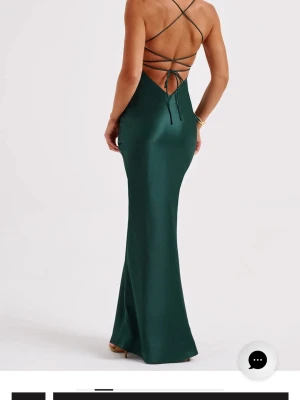 Balklänning  - Elegant mörkgrön långklänning i satinliknande material med smala axelband och öppen rygg med snörning. Klänningen har figurnära passform och släpper ut lätt nertill för en snygg siluett. Perfekt för dig som vill sticka ut med stilrena detaljer.