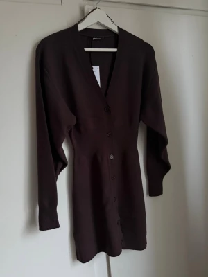 Brun stickad klänning Gina Tricot - Mörkbrun stickad klänning från Gina Tricot med v-ringning och knappar längs hela framsidan. Klänningen har lång ärm och en figurnära passform som ger en snygg siluett. Perfekt för dig som gillar stilrena och enkla plagg.