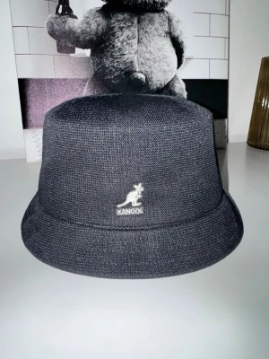 Svart bucket hat från Kangol - Säljer en svart bucket hat från Kangol med det klassiska känguru-logotypen broderad framtill. Hatten är tillverkad i ett lätt och ventilerande material, perfekt för en avslappnad streetwear-look. Modellen heter Tropic Bin och har en mjuk, rundad form.