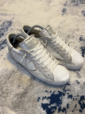 Vita sneakers från Philippe Model - Snygga vita sneakers från Philippe Model med klassisk låg siluett. Skorna har vita skosnören, detaljer i mocka och läder samt märkets logga på sidan. Yttersulan är i gummi och har ett diskret mönster. Perfekta för en clean och stilren look.