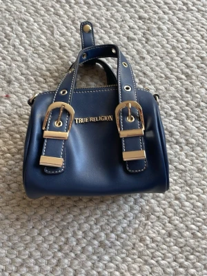 Mörkblå True Religion handväska - KÖPT I NEW YORK OCH NÄSTAN ALDRIG ANVÄND DÄRAV PRIS🤍🤍Snygg mörkblå handväska från True Religion i slätt konstläder med två dekorativa spännen i guld framtill. Väskan har dubbla handtag, vita kontrastsömmar och dragkedja upptill. Perfekt accessoar för att lyfta din outfit. Skriv för fler bilder🤍🤍 