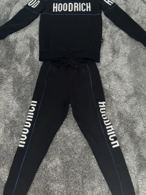 Svart Hoodrich set med tryck XS - Säljer ett svart Hoodrich set med matchande tröja och byxor. Setet har vit HOODRICH-logga tryckt på både tröjan och längs benen. Byxorna har dragsko i midjan och tunna blå sömmar längs sidorna. Perfekt för en streetwear-look.
