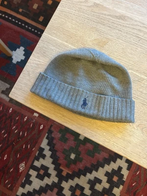 Grå mössa från Polo Ralph Lauren - Snygg grå mössa från Polo Ralph Lauren merinoull. mörkblå logga broderad framtill. Riktigt varm och skön.
