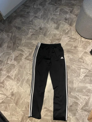 Svarta Adidas mjukisbyxor med vita ränder - Säljer ett par klassiska svarta mjukisbyxor från Adidas med de ikoniska tre vita ränderna längs benen och Adidas-logga på låret. Byxorna har resår i midjan med dragsko och raka ben. Perfekta för chill eller träning.