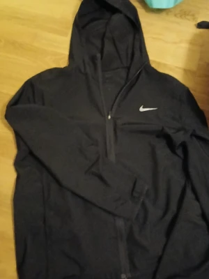 Svart Nike vindjacka med huva - Svart vindjacka från Nike med huva och hel dragkedja framtill. Jackan har en stilren design med Nike-logga på bröstet och är tillverkad i lätt, syntetiskt material som andas. Perfekt för aktiva dagar och enkel att matcha med olika outfits.  /pris kan diskuteras