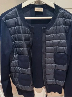 Mörkblå cardiganjacka från Moncler - Snygg mörkblå cardiganjacka från Moncler med quiltade partier framtill och två fickor på varje sida. Jackan har rund halsringning, dragkedja och är tillverkad i en mix av stickat tyg och glansigt material på framsidan. Perfekt för lager på lager och stilren look. Storlek L men mer som M/S.