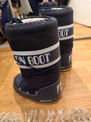 Mörkblå Moon Boot snow boots - Säljer ett par klassiska mörkblå Moon Boot snow boots med vit logga och snörning framtill. Bootsens ovandel är i nylon och de har en vadderad insida för extra värme. Sulan är i gummi och designen är ikonisk och chunky.