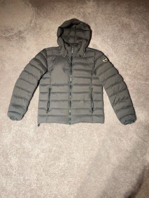 Mörkgrå Colmar jacka   - Snygg mörkgrå dunjacka med huva och två dragkedjeförsedda fickor framtill. Jackan har quiltad design och ett runt märke på ärmen. Perfekt för kalla vinterdagar och ger en clean look. Storlek M känns som S