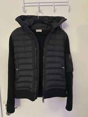 Moncler cardigan - Snygg svart dunväst från Moncler med stickade svarta ärmar och huva. Västen har dragkedja framtill, två fickor med tryckknappar och en extra ficka med dragkedja på ärmen. Perfekt för lager-på-lager och streetstyle.