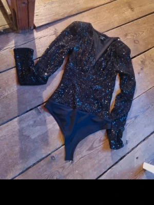Svart glittrig body med paljetter - Snygg svart body med långa ärmar och djup v-ringning. Hela ovandelen är täckt av svarta paljetter som ger ett glittrande intryck. Axelpartiet är lätt puffat för extra attityd. Perfekt för dig som vill sticka ut! Materialet känns stretchigt och bekvämt.