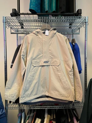 Beige vindjacka från Palace - Snygg beige vindjacka från Palace med stor ficka framtill och huva. Jackan har logga på bröstet och stor PALACE-text på ryggen. Oversized passform och dragkedja vid halsen. Perfekt för streetwear-stil.