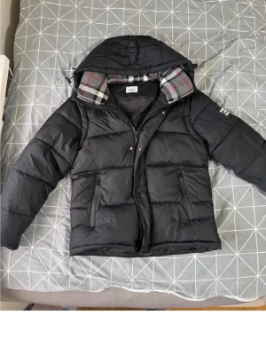 Burberry jacka - Snygg svart dunjacka med avtagbar huva och rutigt foder i grått och rött. Jackan har två stora fickor framtill med dragkedja och tryckknappar, samt diskreta detaljer på ärmen. Perfekt för kalla vinterdagar. Pris kan diskuteras vid snabb affär