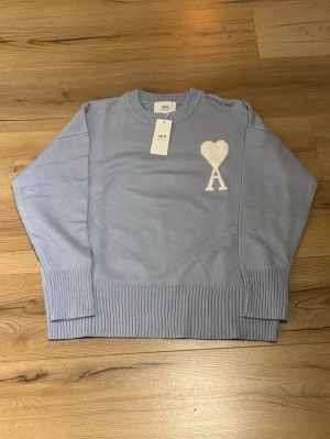 Ljusblå stickad tröja från Ami Paris - Ljusblå stickad tröja från Ami Paris med rund halsringning och ribbade muddar. På bröstet finns ett vitt hjärta med bokstaven 'A' broderat. Tröjan har en avslappnad passform och är perfekt för en trendig vardagslook.