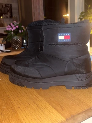 Svarta moon boots från Tommy Jeans - Säljer ett par svarta moon boots från Tommy Jeans med grov sula och vadderat nylonmaterial. Bootsens skaft har dragsko upptill och klassisk Tommy Jeans-logga på sidan. Perfekta för kalla dagar när du vill hålla stilen och värmen.