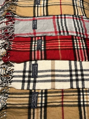 Burberrys rutiga halsdukar i ull - Flera klassiska halsdukar från Burberrys i olika färger: beige, grå, röd och vit. Alla har det ikoniska rutmönstret och fransar i ändarna. Tillverkade i mjuk ull och perfekta för att lyfta din outfit med en tidlös accessoar. Längd mellan 150-180cm autentisisering i pm 100% äkta. 1000kr styck alla färger kostar samma 