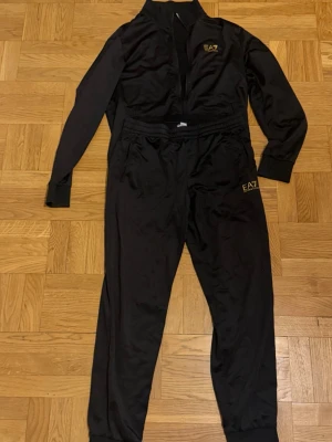 Svart EA7 loungewear set - Snyggt svart loungewear set från EA7 med gul logga på både jacka och byxor. Setet har dragkedja framtill på jackan, muddar vid ärmslut och benslut samt två sidofickor på jackan. Perfekt för chill dagar hemma eller när du vill vara bekväm men ändå se stilren ut.