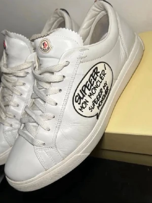 Vita Moncler sneakers - Snygga vita sneakers från Moncler. Jäkligt unika och ovanliga. Skorna har en låg profil, vita skosnören och en unik svart textdetalj på sidan. Tillverkade i slätt läder med klassisk gummisula och Moncler-logga på plösen.