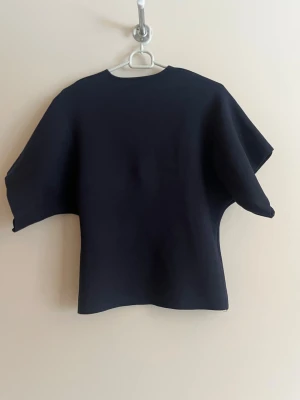 Svart kortärmad kofta med knappar - Stilren svart kofta med korta, vida ärmar och djup V-ringning. Koftan har två dekorativa guldknappar framtill och är tillverkad i ett tjockare, strukturerat material som ger en cool siluett. Perfekt för dig som gillar minimalistisk och modern stil.