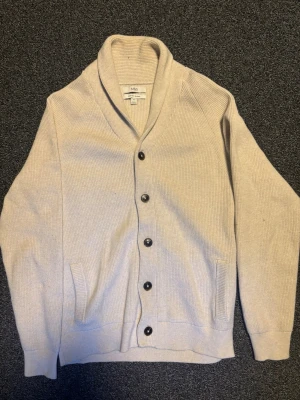 Cardigan - Stilren beige cardigan från M&S i bomullsblandning. Koftan har sjalkrage, ribbstickad struktur, svarta knappar framtill och två fickor.