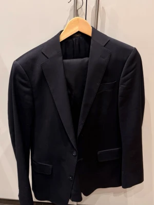 Oscar Jacobsson kostym  - Snygg mörkblå Oscarjacobsson kavaj med två knappar framtill, klassiska slag och tre fickor. Kavajen har en stilren och tidlös design, helfodrad insida och är perfekt för en dressad look. Passar till både skjorta och t-shirt för en modern stil. Storlek 46 på både byxor och kavaj. Skriv gärna om ni vill ha fler bilder. 