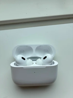 Apple AirPods Pro 2 - Säljer ett par vita Apple AirPods Pro med trådlöst laddningsetui. Hörlurarna har en stilren och kompakt design i plast och levereras med originalkartong, laddningskabel och manualer. Perfekta för dig som vill ha grymt ljud och smidig anslutning till din iPhone.