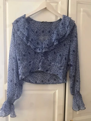 Blå mönstrad blus från Zara - Snygg blå blus från Zara med unikt grafiskt mönster. Blusen har volangkrage, långa ärmar med volang vid ärmslut och är tillverkad i ett tunt, lätt material som ger en luftig känsla. Perfekt för dig som gillar trendiga detaljer och bohemisk stil.