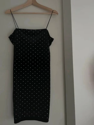 Svart prickig slipklänning - Svart midilång klänning med tunna axelband och vita pärlor över hela tyget. Klänningen har en rak passform. 