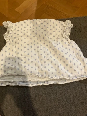 Vit blus med blått mönster och volanger - Söt vit blus med små blå mönster och volangdetaljer vid ärmarna. Blusen har v-ringning och är gjord i ett tunt, luftigt tyg som känns perfekt för sommaren. Den har en avslappnad passform och romantisk känsla.