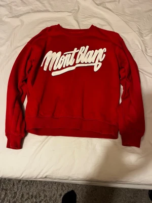 Röd Montblanc sweatshirt från carlings - Snygg röd sweatshirt från Montblanc med vit logga tryckt stort över bröstet. Tröjan har rund halsringning, långa ärmar och ribbade muddar vid ärmslut och nederkant. Perfekt för en avslappnad och trendig look.