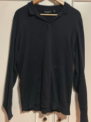 Svart långärmad tröja Brave Soul - Klassisk svart långärmad tröja från Brave Soul med krage och knappdetalj vid halsen. Tröjan har en enkel och stilren design, perfekt för dig som gillar en clean look. Tillverkad i mjukt material för skön känsla.