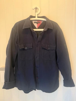 Tommy Hilfiger overshirt - Stilren mörkblå overshirt från Tommy Hilfiger med två bröstfickor med lock och knappar. Skjortjackan har klassisk krage, raka ärmar och knäppning framtill. Perfekt lager-på-lager-plagg i ett mjukt, tjockare tyg som ger en avslappnad look. I storlek L. Hör av er om fler bilder eller funderingar.