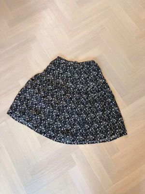 Svart blommig kjol med resårmidja - Söt svart kjol med små vita blommor och resår i midjan. Kjolen har en lätt utställd form och slutar vid knäna. Perfekt för dig som gillar en avslappnad men ändå trendig stil.