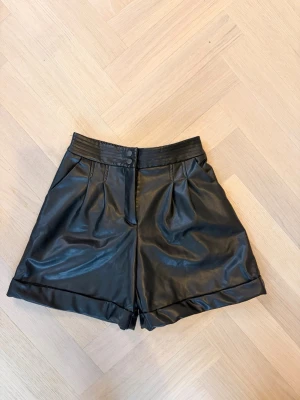 Svarta högmidjade shorts i skinnimitation - Snygga svarta högmidjade shorts i skinnimitation med uppvikta benslut och knappstängning fram. Shortsen har fickor på sidorna och resår baktill för extra komfort. Perfekta för en trendig och edgy look. De är i barnstorlek 140.