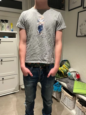 Grå Polo Bear t-shirt från Ralph Lauren - Säljer en grå t-shirt från Ralph Lauren med den ikoniska Polo Bear-trycket på bröstet. T-shirten har korta ärmar, rund hals och är gjord i mjuk bomull. Perfekt för dig som gillar streetstyle och vill ha något klassiskt men ändå lekfullt. Det står att den är storlek M men passar som en s eller xs