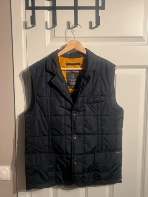 Marinblå dunväst från Oscar Jacobson - Snygg marinblå dunväst från Oscar Jacobson med gul insida och klassisk krage. Västen har knappar framtill, två stora fickor och quiltad design. Perfekt för lager-på-lager och ger en clean look till din outfit.